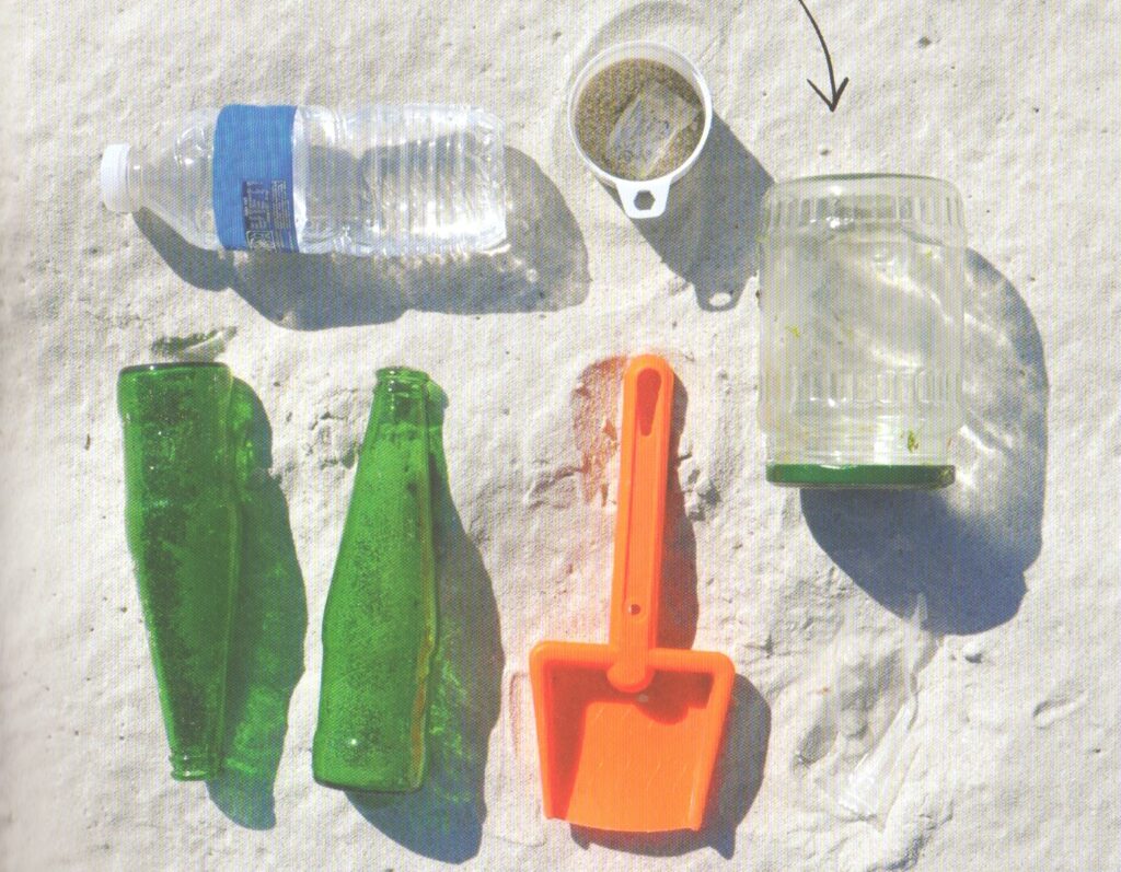 Erforsche das Meer - Flaschen, Becher, Schippe und Glass im Sand drapiert
